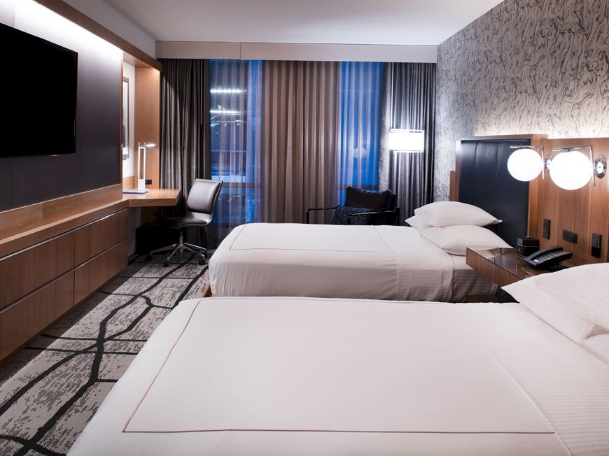 A modern hotel room with two beds, a TV, a desk with a chair, and a decorated wall, with lights and a window with curtains.