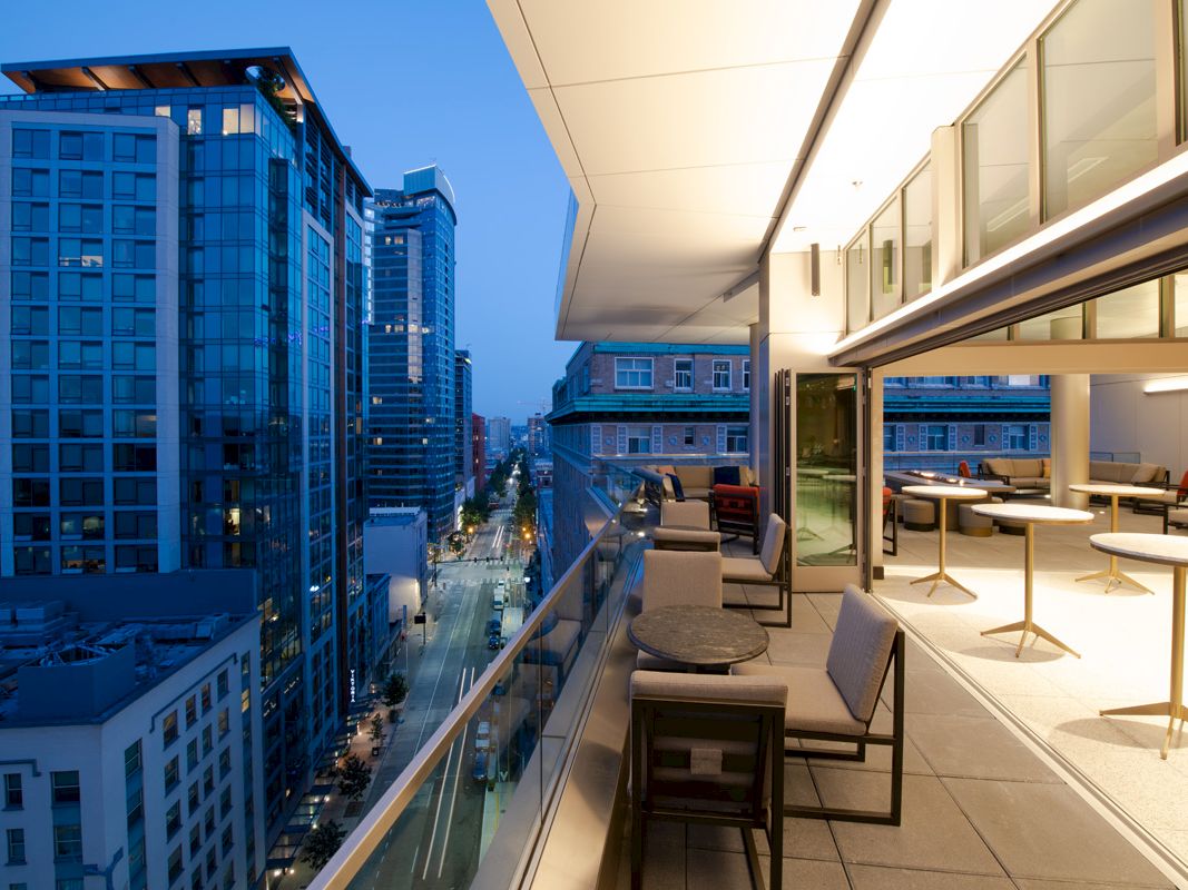 A modern urban balcony with seating overlooks a city street lined with high-rise buildings in the evening.