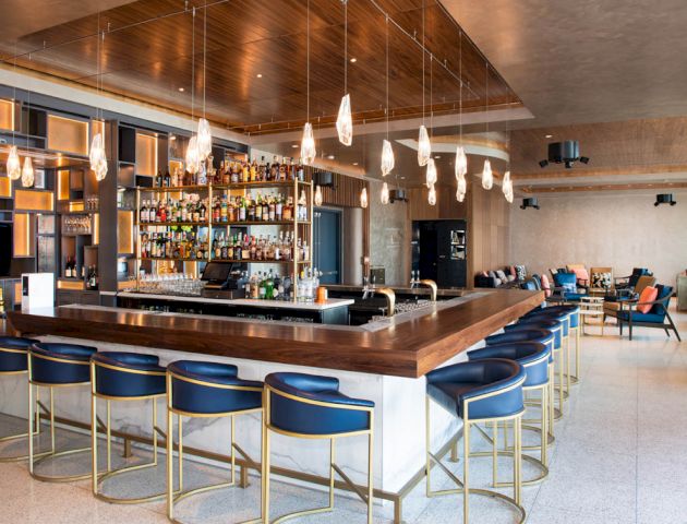 A modern bar with a wooden and metallic design, featuring blue stools, hanging lights, and a well-stocked liquor shelf.