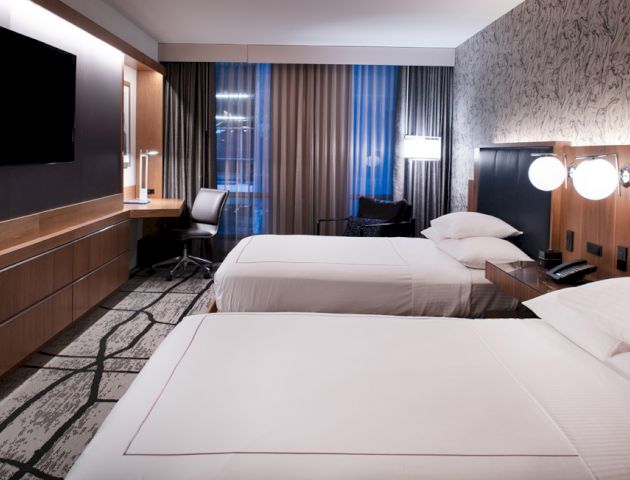 A modern hotel room with two beds, a wall-mounted TV, a work desk, and a window with curtains.