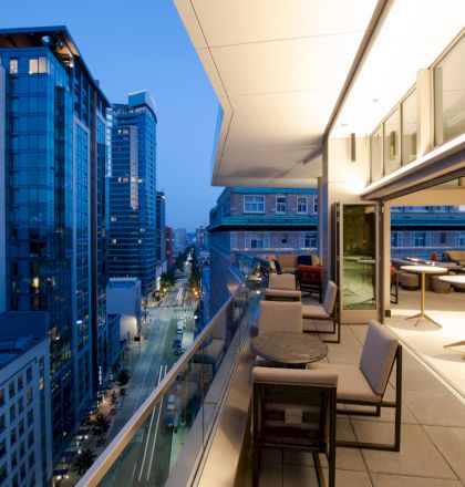 The image shows a modern cityscape with tall buildings during the evening and a terrace with tables and chairs overlooking the street.