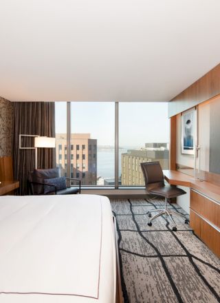 The image shows a modern hotel room with a large bed, wall-mounted TV, work desk, chair, and a window with a city view.