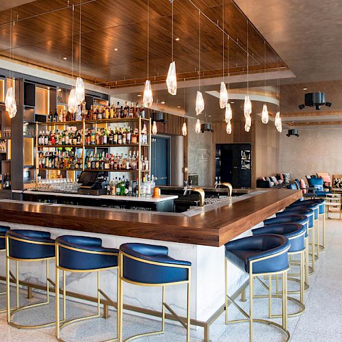 A modern bar with a wooden counter, blue bar stools, numerous hanging lights, and a well-stocked liquor shelf is visible along with seating areas.