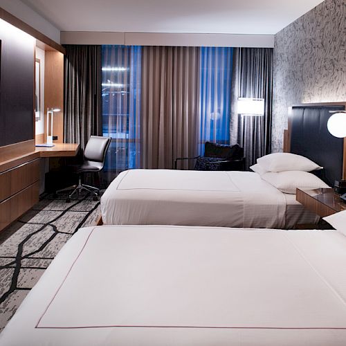 A modern hotel room with two neatly made beds, a TV, a desk with a chair, and a large window with curtains.