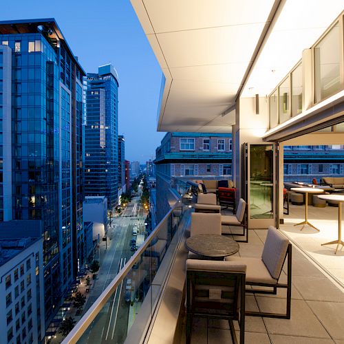 An urban balcony at twilight, with modern furniture and tall buildings lining a street. The balcony features tables and chairs.
