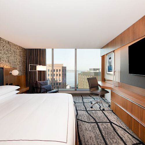 A modern hotel room with a large bed, wall-mounted TV, desk, and chair, featuring a city view through large windows.