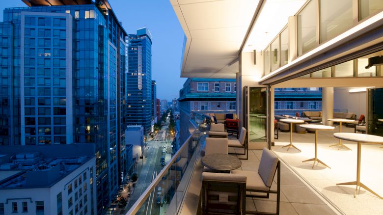 The image shows a modern city view from an outdoor balcony at dusk. The balcony has tables and chairs, and the surrounding buildings are illuminated.