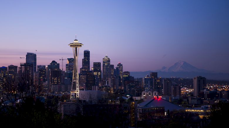 A city skyline at dusk includes a prominent tower, with a mountain in the background and city lights beginning to illuminate.