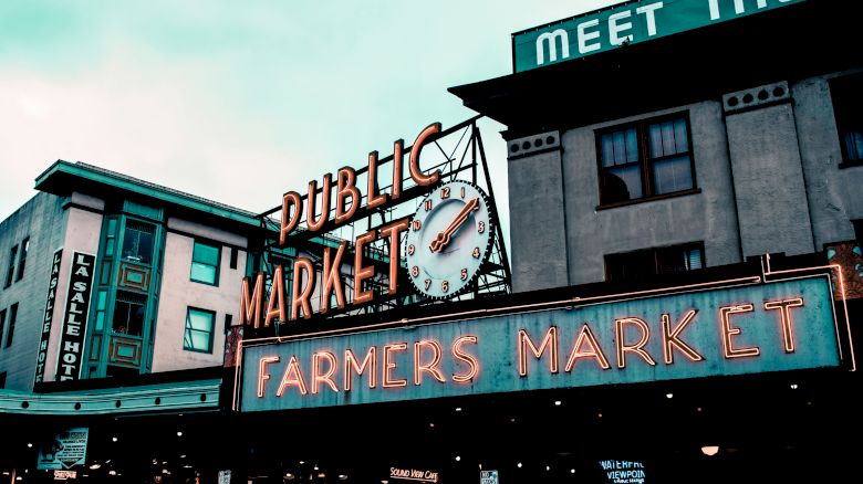 This image shows the iconic Pike Place Market sign in Seattle, with "Public Market" and "Farmers Market" lit up in neon colors, along with surrounding buildings.