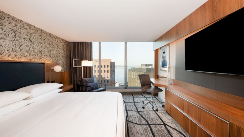 A modern hotel room with a bed, desk, chair, large TV, and cityscape view through a large window. The room is stylish and minimalist.