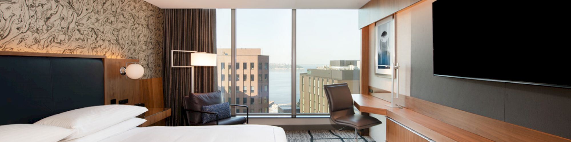 A modern hotel room features a large bed, flat-screen TV, desk, chair, and window with a city view.