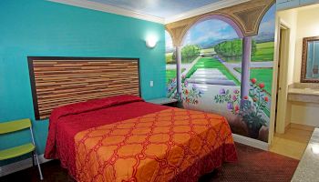 This image shows a colorful hotel room with a decorative mural, a bed with a red and orange cover, a small seating area, and a sink near the bathroom.