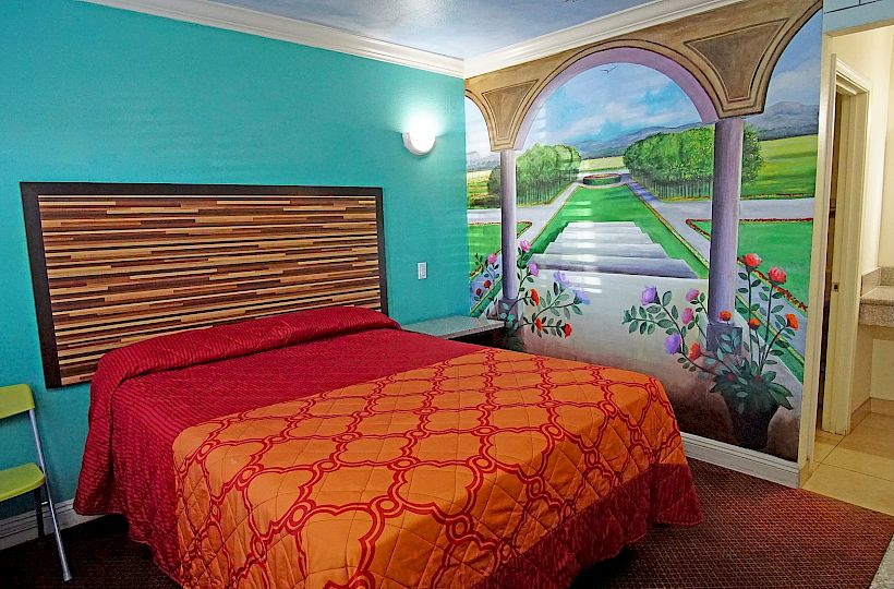 This image shows a colorful hotel room with a decorative mural, a bed with a red and orange cover, a small seating area, and a sink near the bathroom.