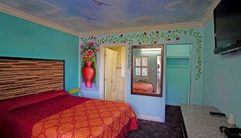 The image depicts a colorful bedroom with a double bed, vibrant wall art, a mirror, and a TV mounted on the wall.