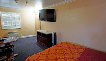 The image shows a simple, well-lit room with a bed, a wall-mounted TV, a table with chairs, and a window with shutters, creating a cozy atmosphere.