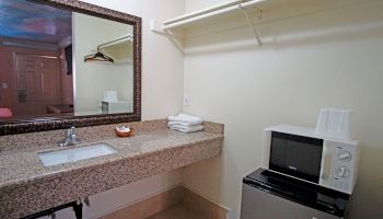 The image shows a small hotel room area with a sink, mirror, microwave, refrigerator, hanging clothes, and a few towels on the countertop.