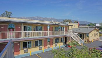 The image shows a two-story motel with exterior entrances to rooms, a staircase, potted plants, and a mountain range in the background.