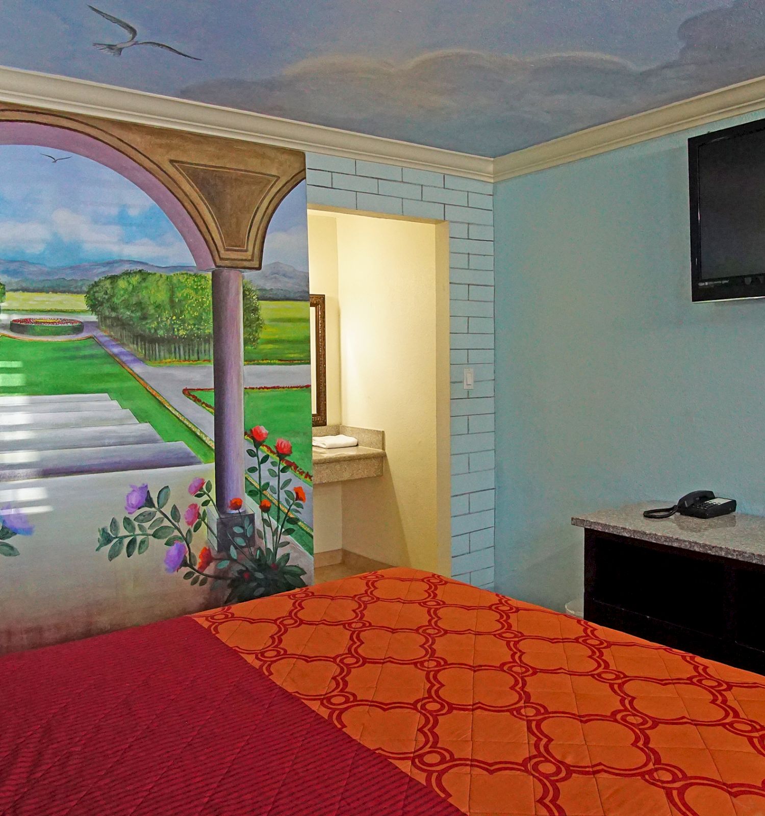 A hotel room with a vibrant wall mural, a large TV, a bed with red and orange linens, and a small vanity area visible through an open doorway.