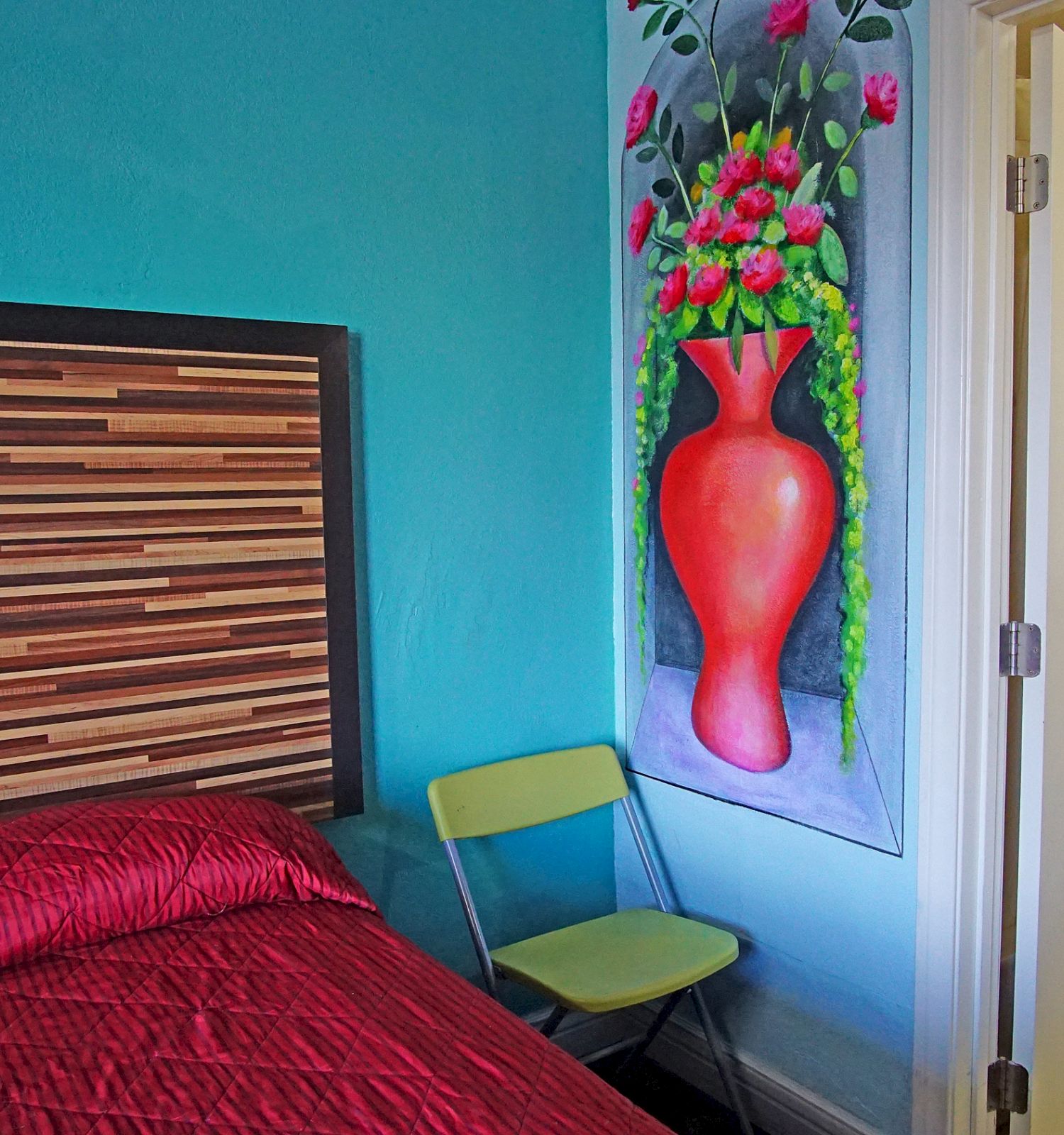 A bedroom with a striped headboard, red bedding, a green chair, floral painting, and an open door to a tiled bathroom with a towel on the rack.