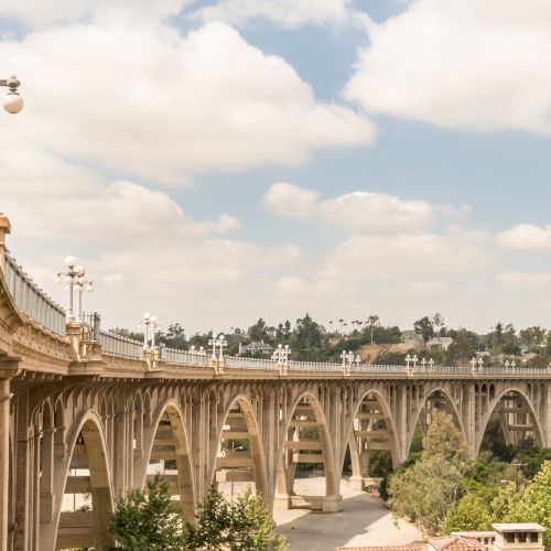 This image shows a large, arched bridge spanning over a road with trees and greenery surrounding it, under a partly cloudy sky.