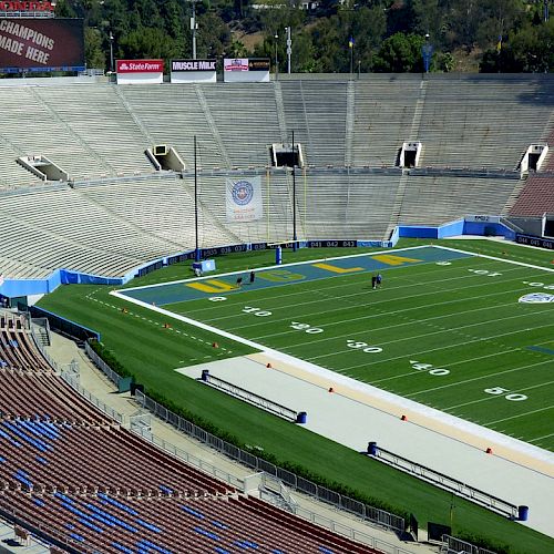 An empty football stadium with a green field, seating areas, and a scoreboard. The scoreboard appears to display a message or announcement.