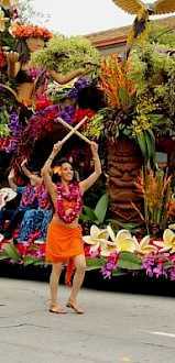 A person in colorful attire dances with sticks in front of a vibrant, flower-decorated float, part of a procession or parade.